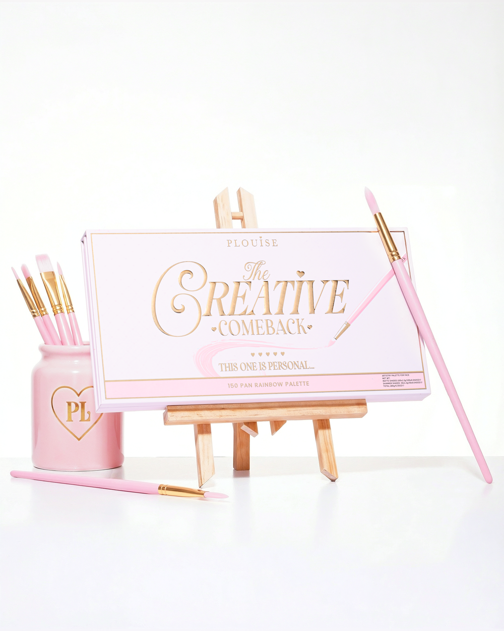 Eine geschlossene rosa P.Louise Creative Comeback XXXL Rainbow-Lidschattenpalette auf einer Kunststaffelei, umgeben von Make-up-Pinseln
