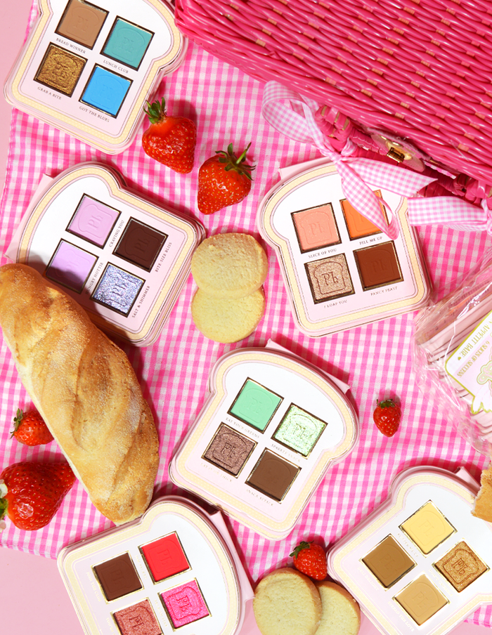 Ein Satz Mini-Lidschattenpaletten im Sandwich-Stil mit verschiedenen Make-up-Farbtönen auf einer rosa karierten Oberfläche, begleitet von Erdbeeren und einem Stück Brot.