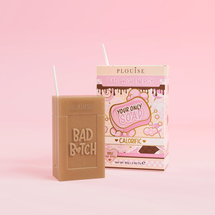 Ein braunes Stück Seife mit der Gravur „Bad Bitch“, begleitet von einer bunten Saftschachtel mit der Aufschrift „Calorific“, präsentiert auf einem rosa Hintergrund.