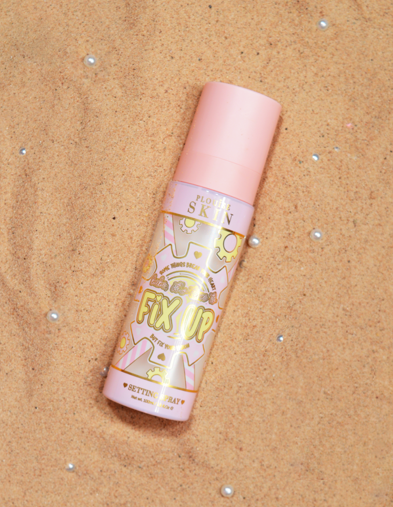 Eine Flasche P.Louise Fix Up Mattifying Setting Spray mit rosa Verschluss und dekorativen Mustern auf dem Etikett, ausgestellt auf einer sandigen Oberfläche.
