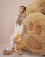 Frau im Teddy-Pyjama hält einen Teddybären neben einer großen Plüschbärentatze auf beigem Hintergrund