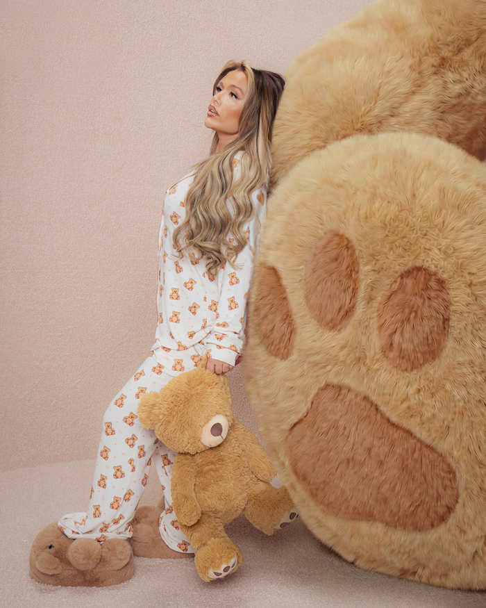Frau im Teddy-Pyjama hält einen Teddybären neben einer großen Plüschbärentatze auf beigem Hintergrund